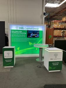 RENTAL: Modified RE-1081 Design with White Laminated Backwall with Recessed Single-Sided Lightbox, RE-1575 Reception Counter, Workstation Pedestal, RE-704 Charging Station Table, Large Monitor Mount, 43" Monitor, SEG Backlit Fabric Graphic, Vinyl Applied